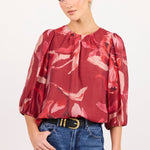 A woman modeling a red and pink floral print blouse with puff sleeves, paired with blue jeans and a black belt, against a plain white studio background.