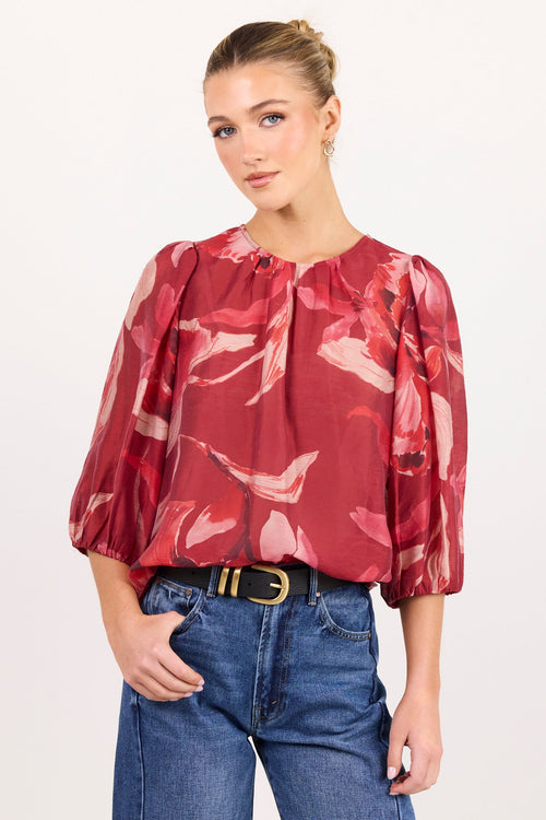 A woman modeling a red and pink floral print blouse with puff sleeves, paired with blue jeans and a black belt, against a plain white studio background.