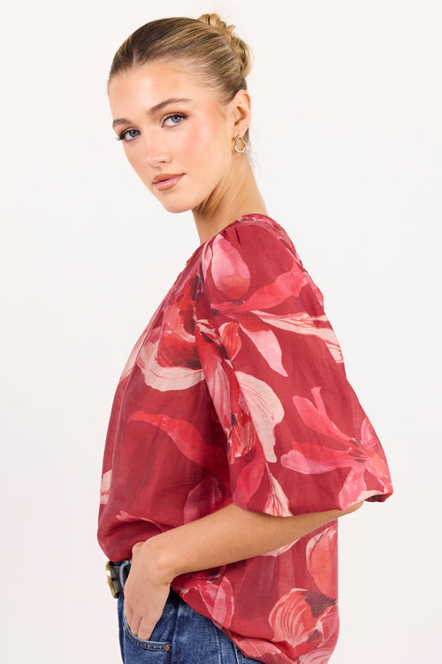 A woman modeling a red floral chiffon blouse with puff sleeves and blue jeans against a plain white studio background.