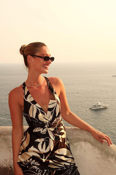 model wears Front-facing view of slim oval sunglasses in a glossy black frame with smoke-tinted lenses. The sleek, narrow silhouette channels a strong 90s influence, offering a bold, minimalist look.