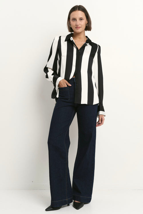 Woman wearing a black and white vertically striped button-up shirt with dark denim trousers, posed casually against a plain wall.