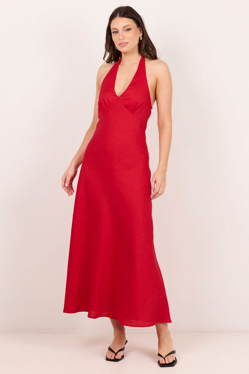 A woman modeling a red halter-neck maxi dress with a plunging V-neckline, standing against a plain off-white studio background.
