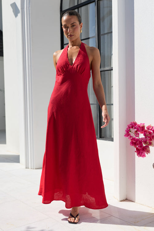 A woman modeling a sleeveless, halter-neck, floor-length red dress in an outdoor setting with white columns and a potted pink orchid.