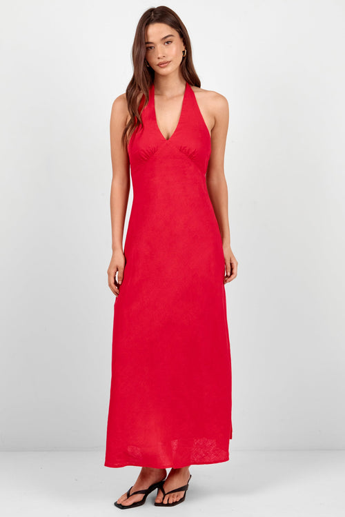 A woman modeling a sleeveless, halter-neck maxi dress in a vibrant red color, standing against a plain light gray studio background.