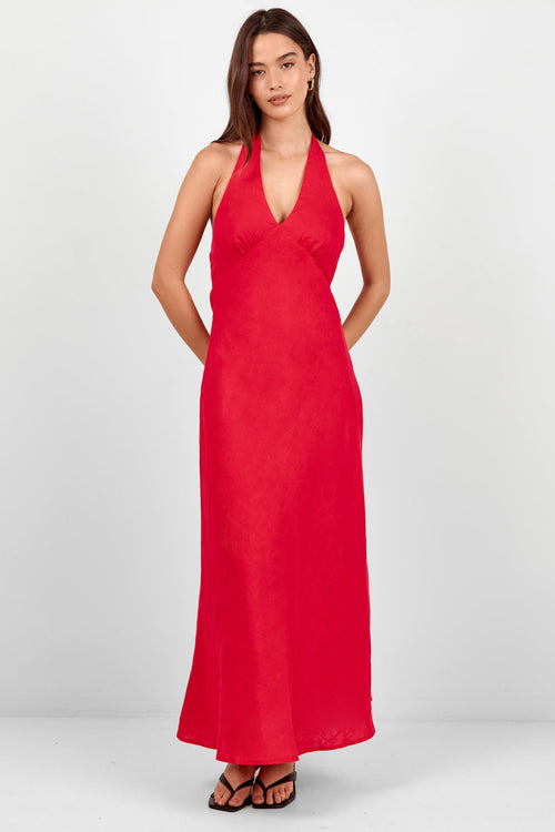 A woman modeling a bright red halter-neck maxi dress with a deep V-neckline and side pockets, paired with black strappy sandals, against a light gray studio background.