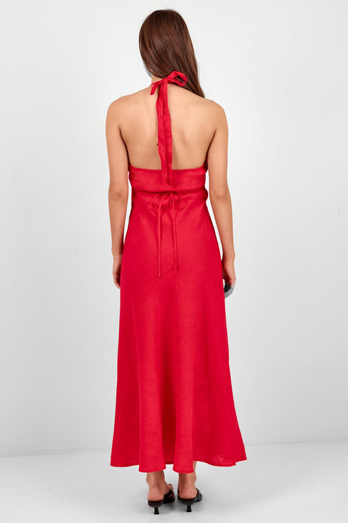 A woman viewed from the back wearing a bright red halter-neck maxi dress with a tie at the back and black heels, standing against a plain light gray studio background.