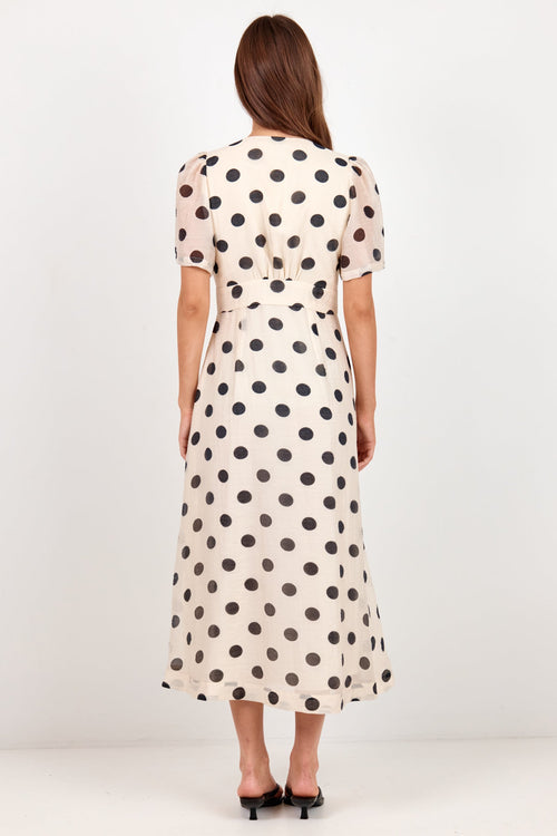 A woman viewed from behind wearing a cream-colored midi dress with black polka dots, standing against a plain white studio background.