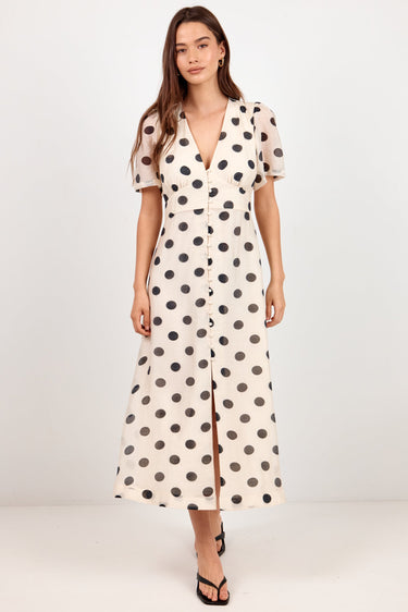 A woman modeling a cream-colored midi dress with black polka dots, featuring short puff sleeves, a V-neckline, and a front slit, set against a plain white studio background.