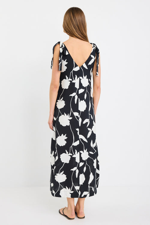 Woman wearing a sleeveless black midi dress with large white floral print, tie details at the shoulders, and a relaxed flowing fit styled with black sandals.