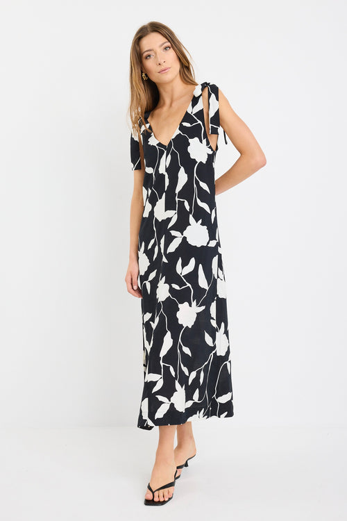Woman wearing a sleeveless black midi dress with large white floral print, tie details at the shoulders, and a relaxed flowing fit styled with black sandals.