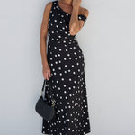 A woman modeling a black sleeveless maxi dress with white polka dots, standing against a minimalist white stucco wall.