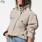 A woman wearing a light beige cropped utility jacket with black buttons, styled with blue jeans and sunglasses, posing against a white textured wall.