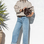 A woman stands outdoors against a white wall wearing a light beige utility jacket, light blue wide-leg jeans, and brown sandals, holding a dark brown handbag.
