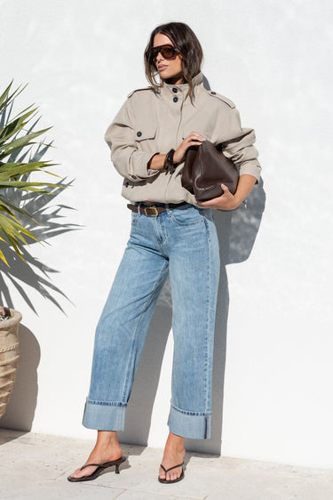 A woman stands outdoors against a white wall wearing a light beige utility jacket, light blue wide-leg jeans, and brown sandals, holding a dark brown handbag.