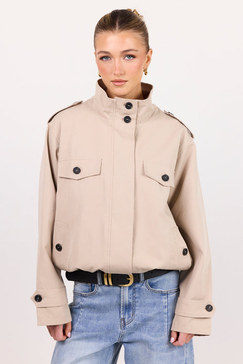 A woman modeling a cropped beige military-style jacket with a high stand collar, flap chest pockets, and black buttons, paired with light blue jeans and a black belt, against a plain off-white studio background.