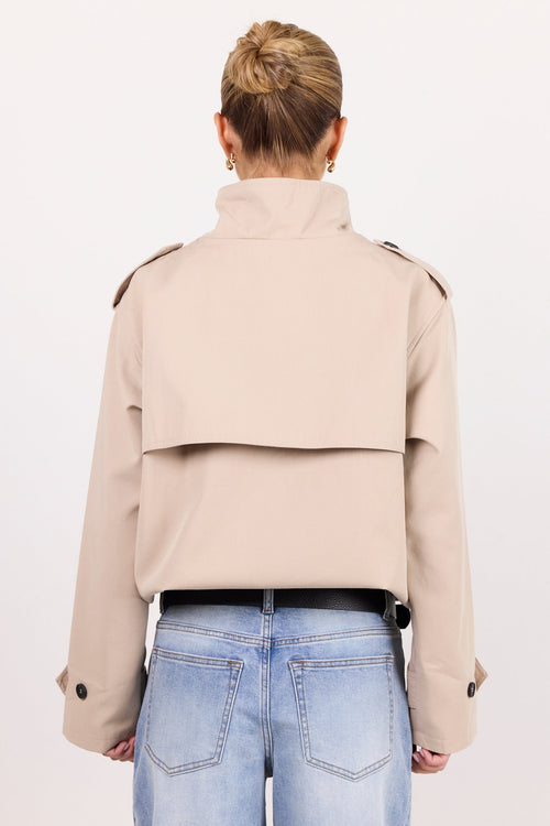 A model viewed from behind wearing a light beige cropped trench jacket with shoulder epaulets, paired with light blue jeans and a black belt, against a plain white studio background.
