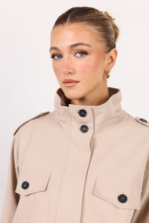A woman wearing a beige military-style jacket with a high stand-up collar and dark buttons, photographed against a clean white studio background.