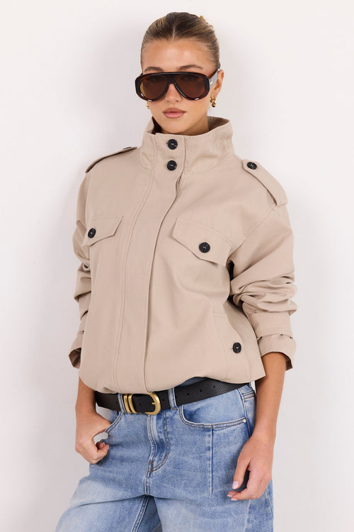 A woman modeling a beige cropped utility jacket with black buttons, paired with light blue jeans and dark sunglasses, against a plain white background.