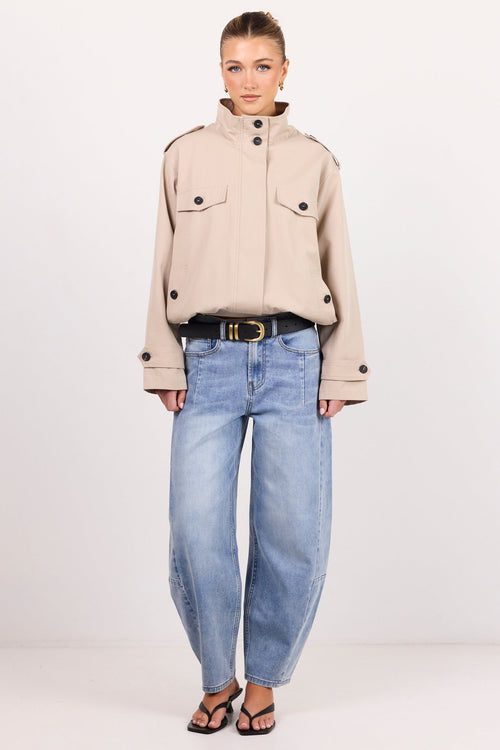 A model stands against a plain white studio background wearing a beige cropped military-style jacket with black buttons, light-wash blue wide-leg jeans, a black belt with a gold buckle, and black sandals.