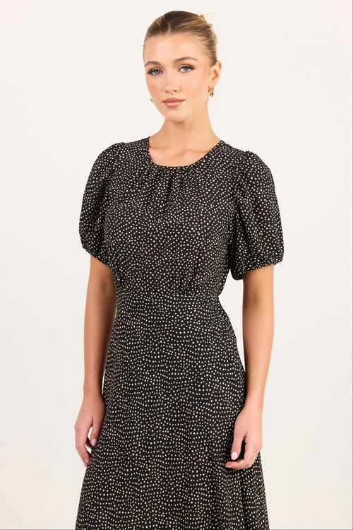 A woman modeling a short-sleeved black dress with a dense white micro-dot pattern, featuring a gathered round neckline and puffed sleeves, set against a plain off-white studio background.
