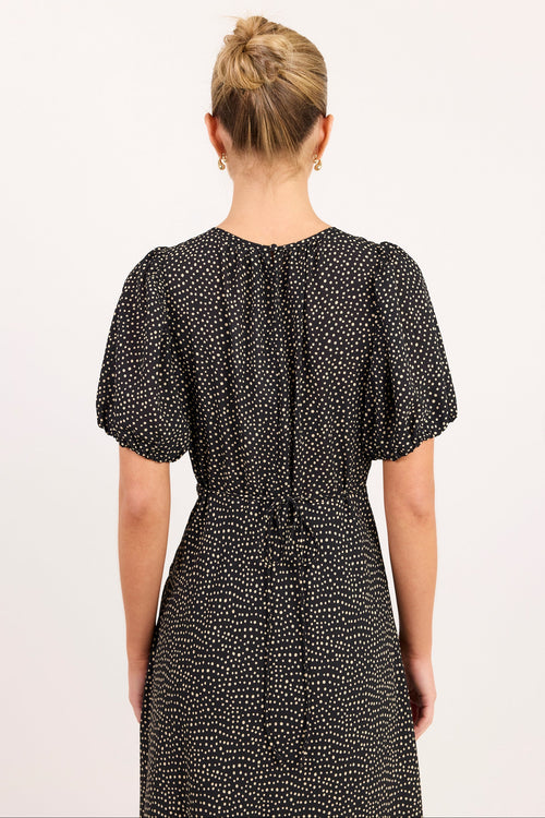 A woman viewed from the back wearing a short-sleeve black dress with a small white allover print, set against a plain off-white background.