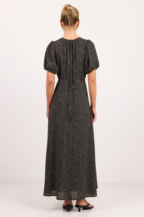 A woman viewed from the back wearing a black midi dress with a small white abstract print, short puff sleeves, and a tie at the back waist, standing against a plain white studio background.