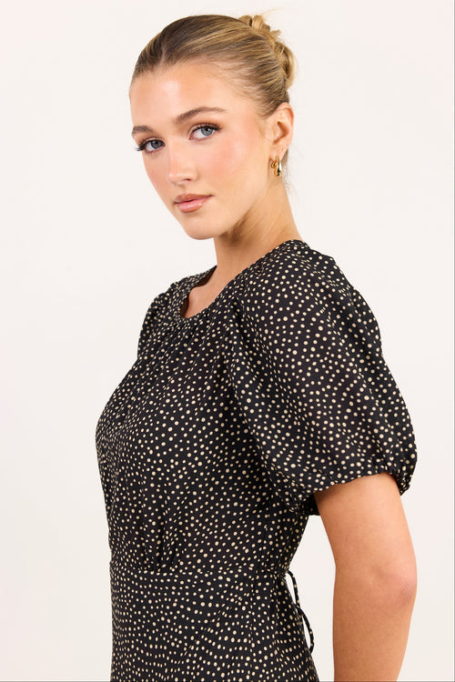 A woman wearing a black short-sleeve top with a white polka dot pattern, shown from a side-back angle against a plain light background.
