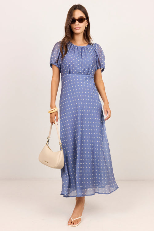 A woman modeling a blue midi dress with white polka dots, featuring short puff sleeves and a gathered neckline. She is holding a cream-colored handbag and wearing sunglasses, set against a plain light beige studio background.