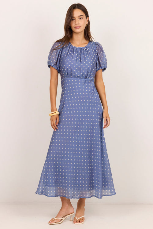 A woman modeling a blue midi dress with a white polka dot pattern, short puff sleeves, and a gathered waist, standing against a plain off-white studio background.