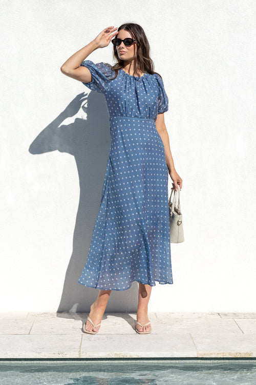 A woman wearing a blue and white patterned midi dress stands next to a swimming pool in front of a white wall, holding a small beige handbag.