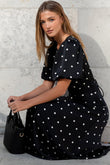 Woman in a black polka dot dress holding a black handbag against a light gray wall.