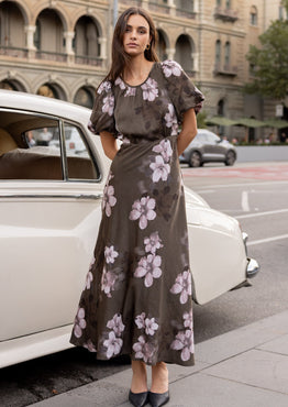 Woman in a floral dress standing next to a vintage car on a city street.