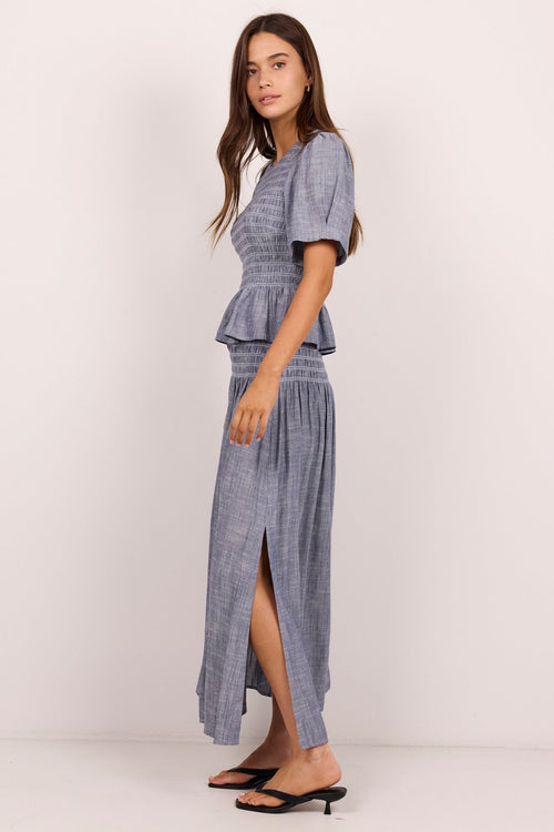 A woman modeling a matching two-piece set consisting of a short-sleeve peplum top and a high-slit maxi skirt in a blue and white textured pattern, paired with black flat sandals, against a plain off-white studio background.
