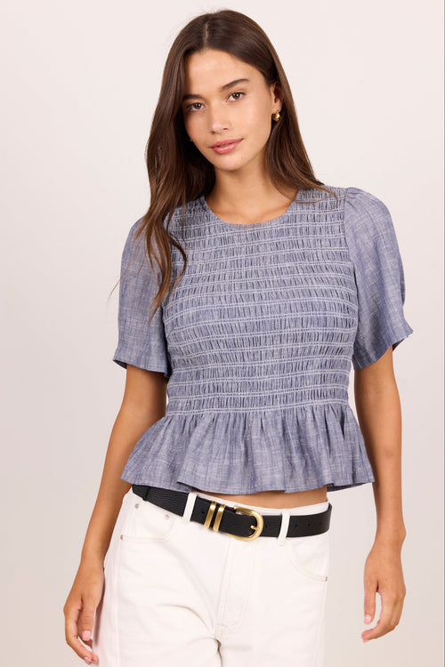 A woman modeling a short-sleeve smocked blouse in a dusty periwinkle blue color, paired with white pants and a black belt, against a plain light beige background.