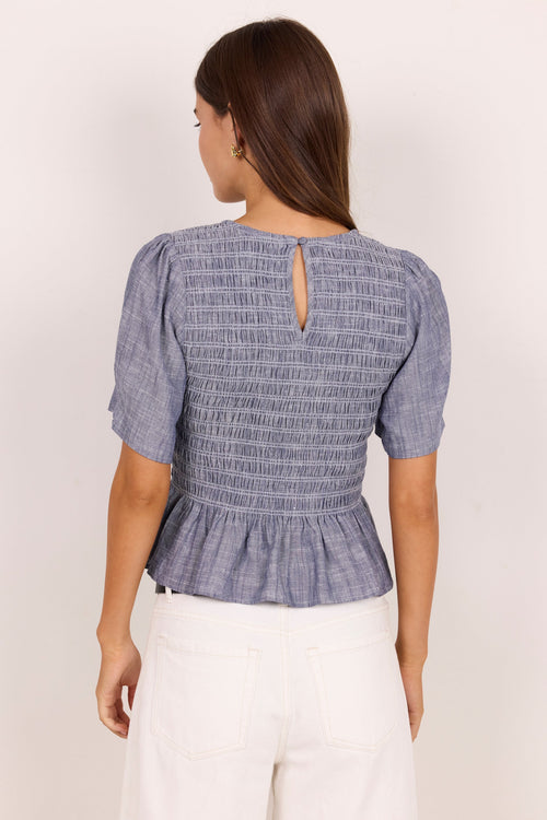 A woman viewed from the back wearing a smocked short-sleeve top in a blue and white textured pattern, paired with white pants, against a plain light background.
