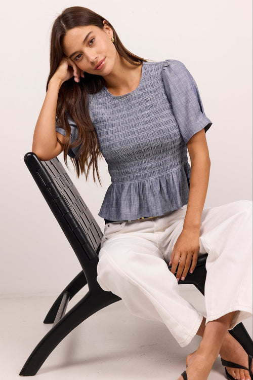 A woman models a blue-gray smocked short-sleeve peplum top paired with white cropped pants and black sandals, seated on a black modern chair against a plain off-white studio background.