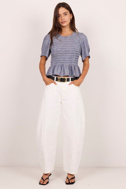 A woman modeling a short-sleeve smocked top in a blue and white pattern, paired with white wide-leg pants and black sandals, standing against a plain off-white studio background.