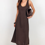 A woman modeling a sleeveless, scoop-neck midi dress in a dark brown color. She is wearing black sunglasses, holding a matching dark brown clutch, and wearing black strappy sandals against a plain white studio background.