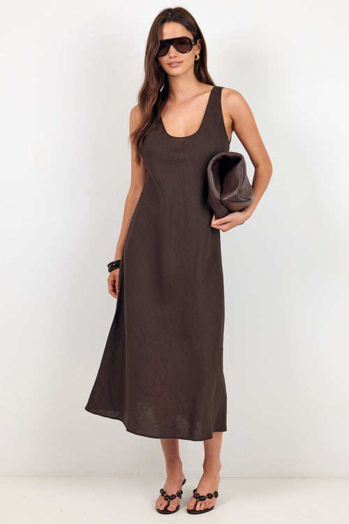A woman modeling a sleeveless, scoop-neck midi dress in a dark brown color. She is wearing black sunglasses, holding a matching dark brown clutch, and wearing black strappy sandals against a plain white studio background.