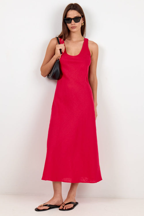 A woman modeling a sleeveless bright red midi dress with a scoop neckline, paired with black sunglasses, a black shoulder bag, and black flip-flop sandals, standing against a plain white studio background.
