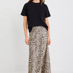 Woman wearing a satin leopard print midi skirt with a bias cut and elastic waist, paired with a black short sleeve tee and black flats.