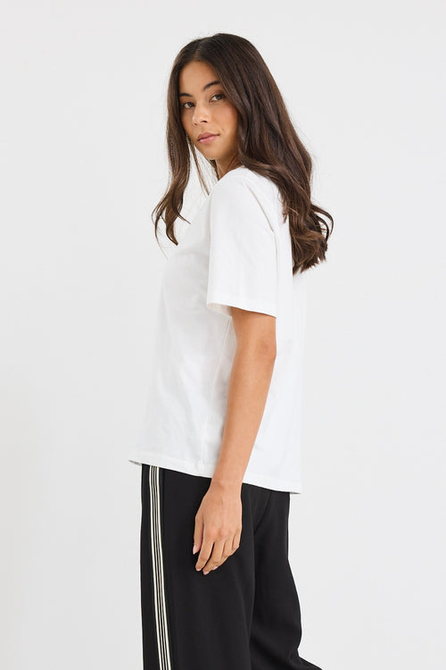 Woman wearing a relaxed-fit ivory short sleeve tee tucked into high-waisted black trousers, styled with minimal makeup and gold earrings.







