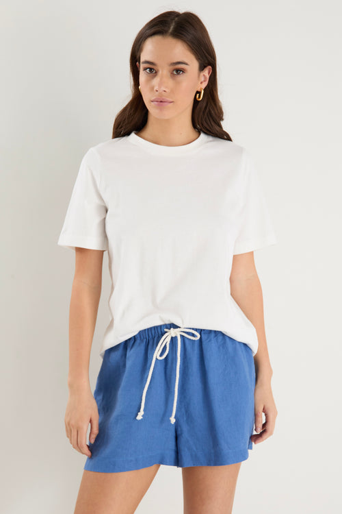 Woman wearing high-rise cornflower blue linen pull-on shorts with rope tie detail, styled with a plain ivory tee.