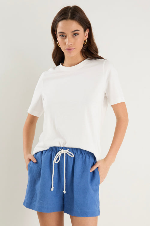 Woman wearing high-rise cornflower blue linen pull-on shorts with rope tie detail, styled with a plain ivory tee.