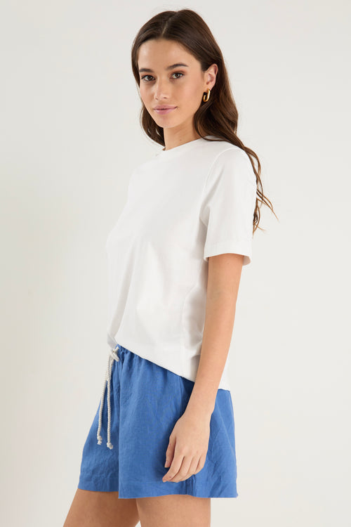 Woman wearing high-rise cornflower blue linen pull-on shorts with rope tie detail, styled with a plain ivory tee.