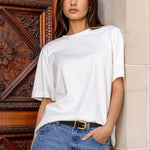 Woman in a white top and blue jeans standing against a stone wall with a wooden door.