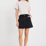 Woman wearing a washed black denim mini skirt with front patch pockets, styled with a tucked-in neutral tee and black sandals.