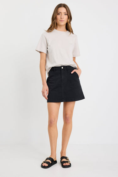 Woman wearing a washed black denim mini skirt with front patch pockets, styled with a tucked-in neutral tee and black sandals.