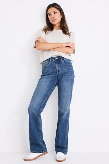 Woman wearing mid-rise, mid blue wash jeans with a plain beige t-shirt and nude sandals.