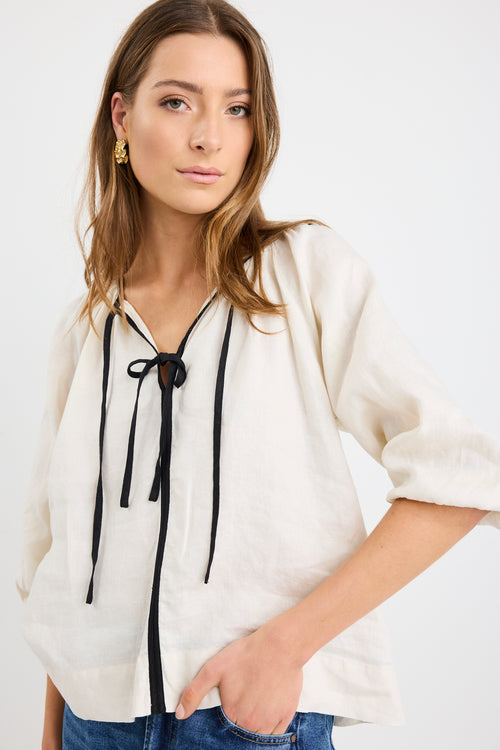 Woman wearing a cream long sleeve linen top with black trim and tie-front detail, designed in an oversized fit with puff sleeves, paired with blue jeans.